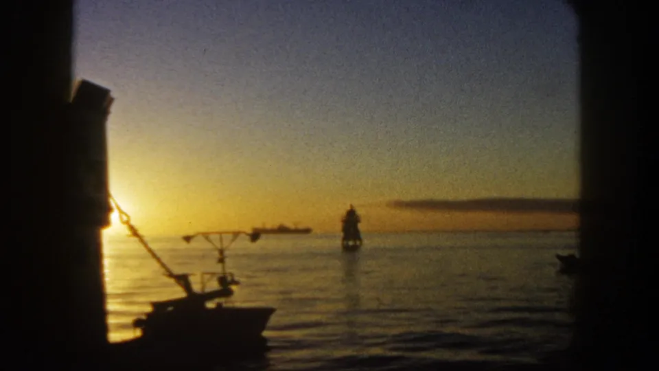 1962 San Diego 8mm frame with the sun near the horizon, a foreground boat silhouette, and a distant lighthouse or navigation marker offshore.