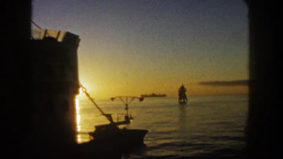 1962 San Diego sunset frame showing a dark foreground boat, calm ocean, distant ship, and offshore lighthouse or navigation marker.