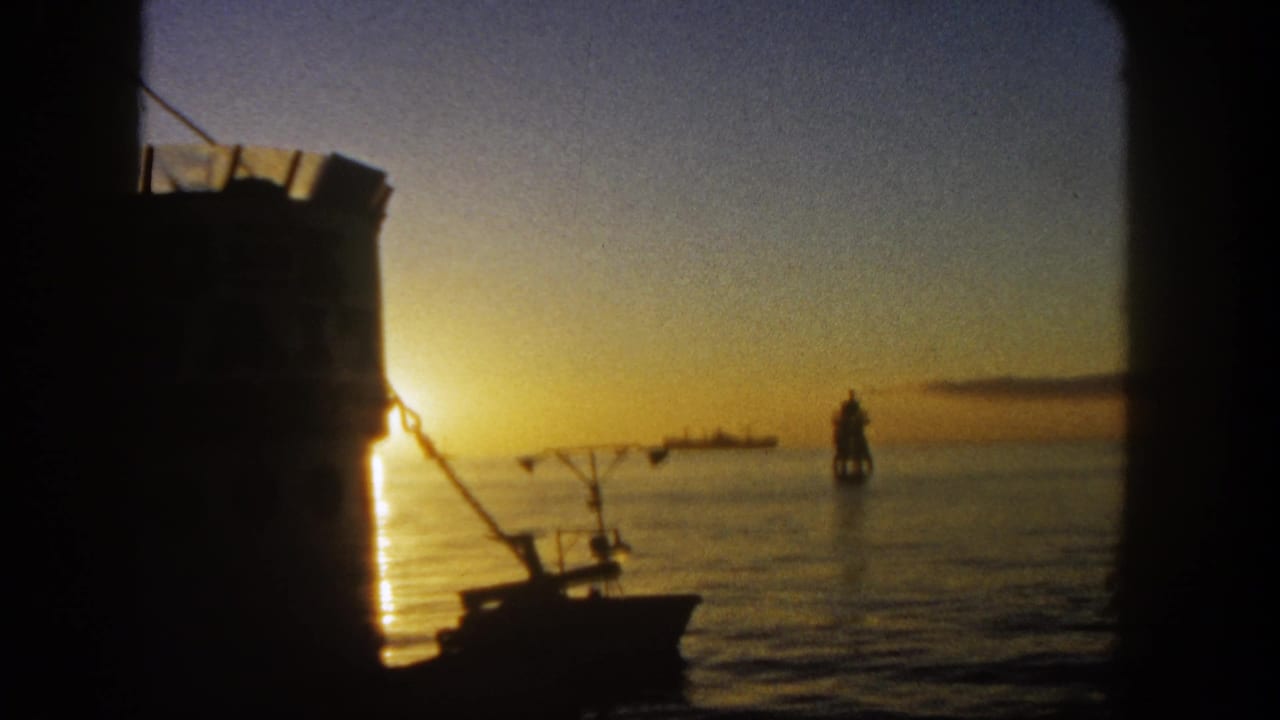 1962 San Diego 8mm frame showing a low sunset over calm harbor water, with a silhouetted boat, rigging, and dark vessel framing.