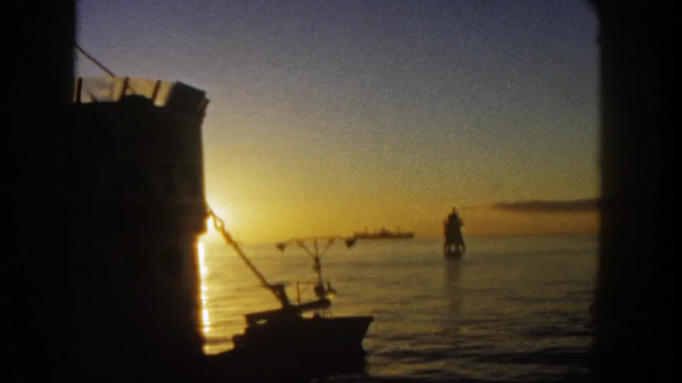 1962 San Diego 8mm frame showing a low sunset over calm harbor water, with a silhouetted boat, rigging, and dark vessel framing.
