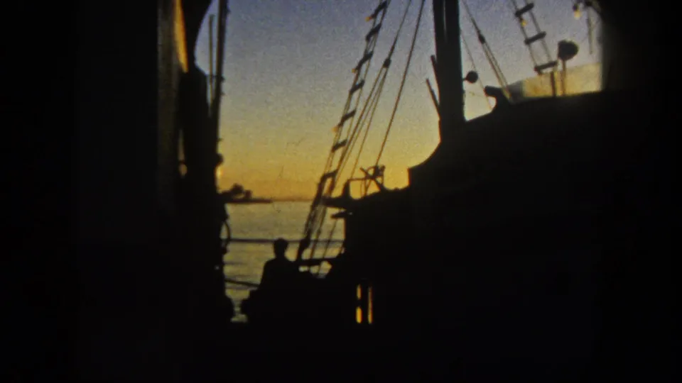 1962 San Diego waterfront frame with dark rigging, a seated or standing silhouetted person, calm water, and sunset color in the distance.