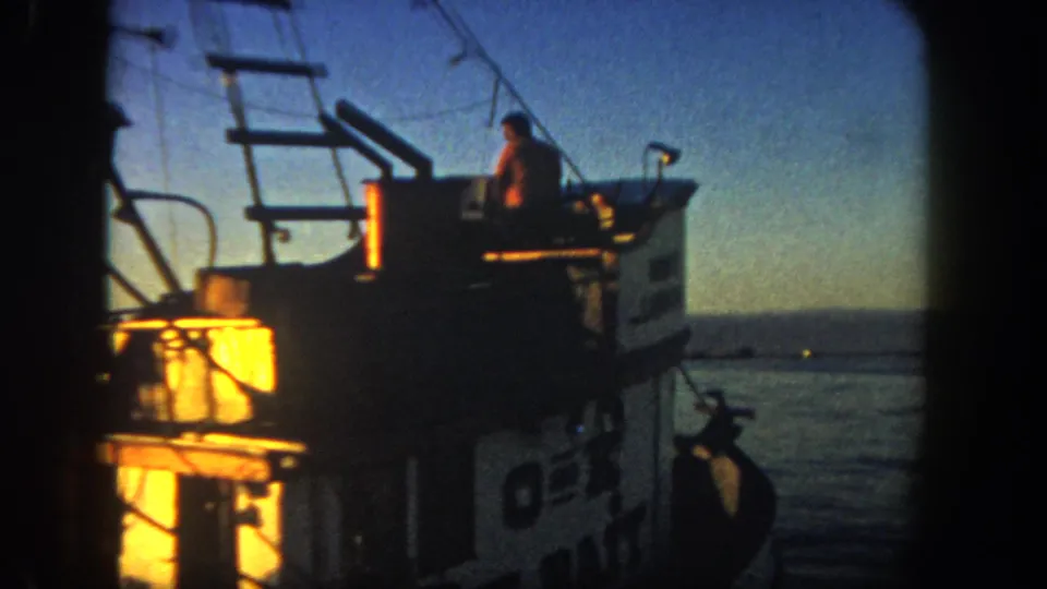 A person stands on a boat deck in the 1962 San Diego harbor, with cabin windows, rails, rigging, and evening water behind them.