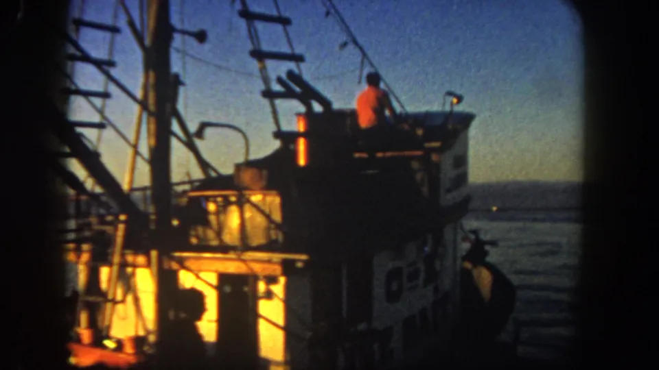 1962 San Diego coastal 8mm frame showing a fishing boat at dusk, with warm cabin light, rigging, ladder, calm water, and a crew member silhouetted on the upper deck.