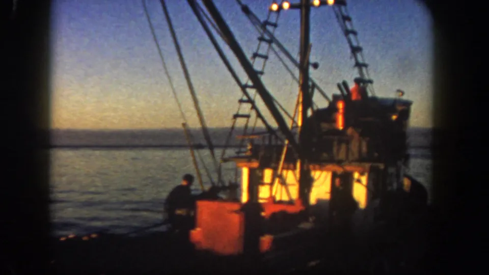1962 8mm frame of a San Diego fishing boat at dusk, showing the lit cabin, rigging, deck equipment, water, horizon, and crew figures partly silhouetted.