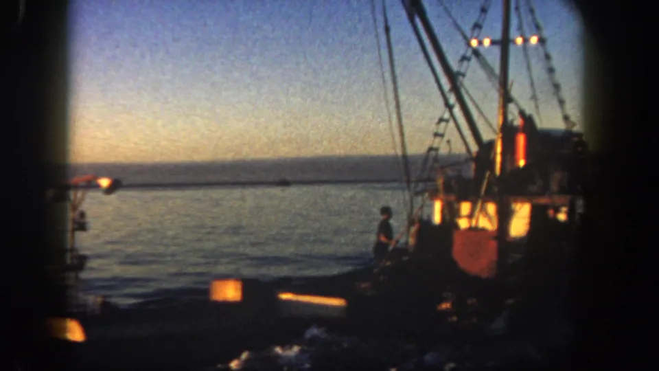 1962 San Diego 8mm frame showing a fishing boat in silhouette at dusk, with rigging, deck lights, a visible person, and dark water in the foreground.