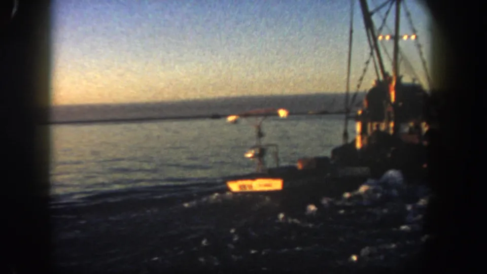 1962 San Diego home-movie frame showing a small lighted vessel being towed behind or alongside a larger fishing boat, with wake and a low horizon at dusk.
