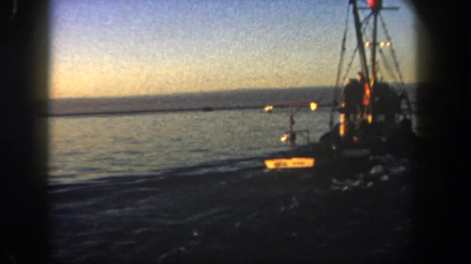1962 San Diego 8mm frame showing fishing boat rigging at right, a small illuminated vessel near center, and calm dark water under a fading sky.
