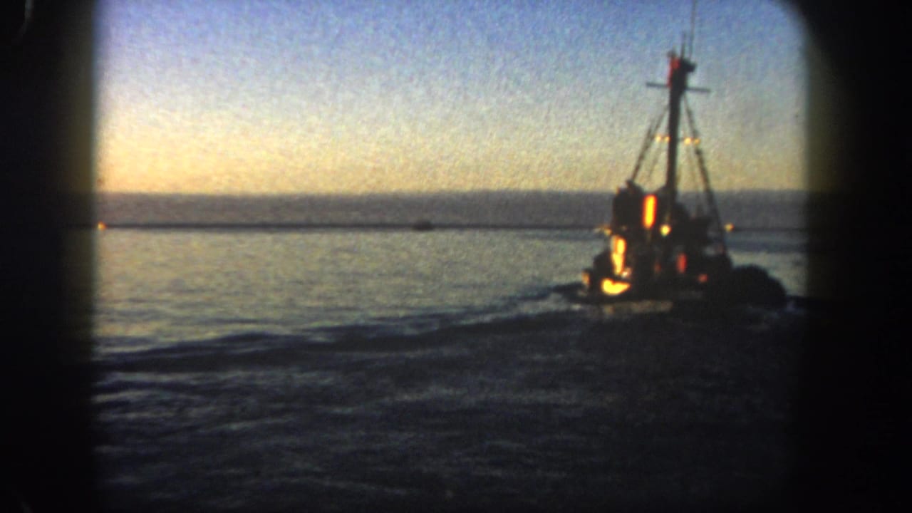 1962 San Diego 8mm frame showing a lit fishing boat on calm harbor water beneath a fading sky.