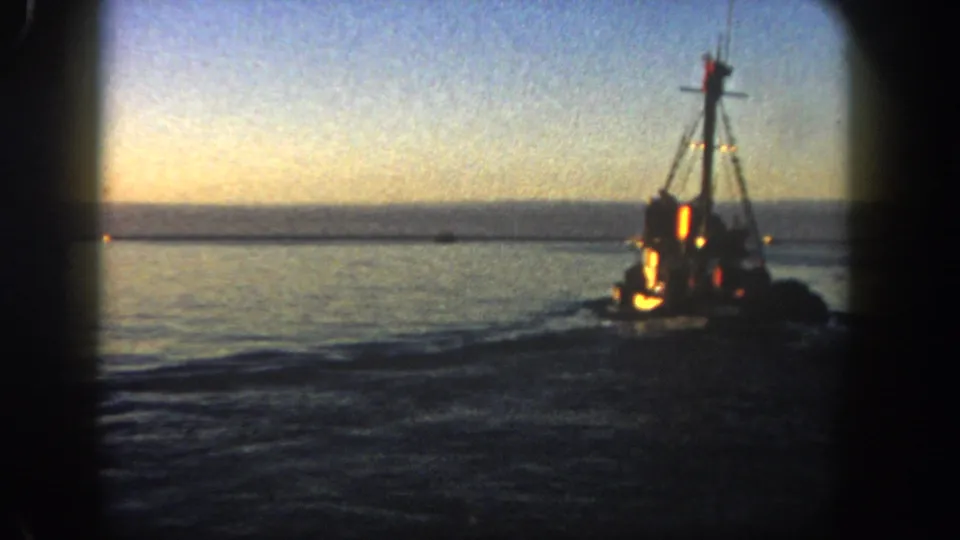 1962 San Diego 8mm frame showing a lit fishing boat on calm harbor water beneath a fading sky.