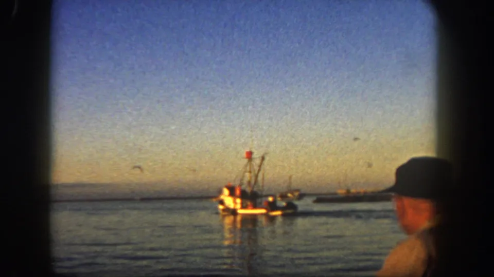 1962 San Diego 8mm frame with a fishing boat offshore and a man in a cap watching from the foreground.