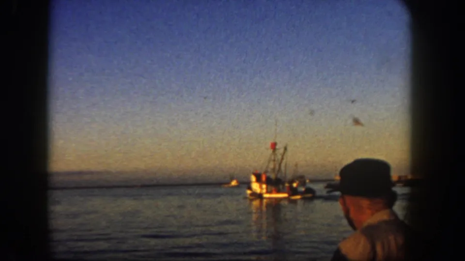 1962 San Diego harbor frame showing a man seen from behind or profile watching a small fishing boat at dusk.