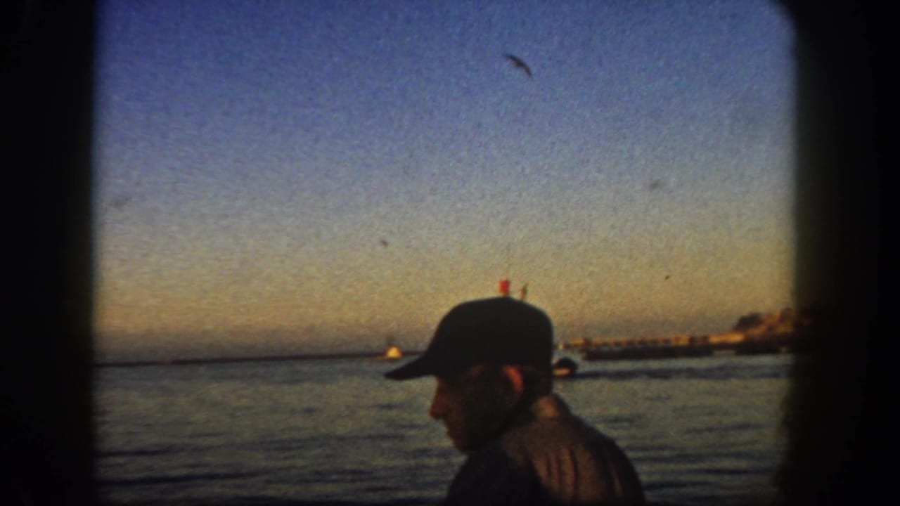 Profile of a man in a cap beside calm water with a boat and shoreline in the distance.