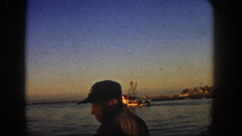 1962 San Diego home-movie view of a capped man near the waterfront, with a small boat and shoreline visible behind him.