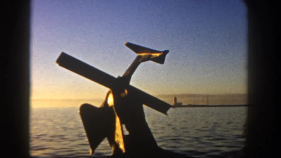 1962 San Diego waterfront frame with a large anchor in silhouette, calm water, and a low sunset horizon behind it.