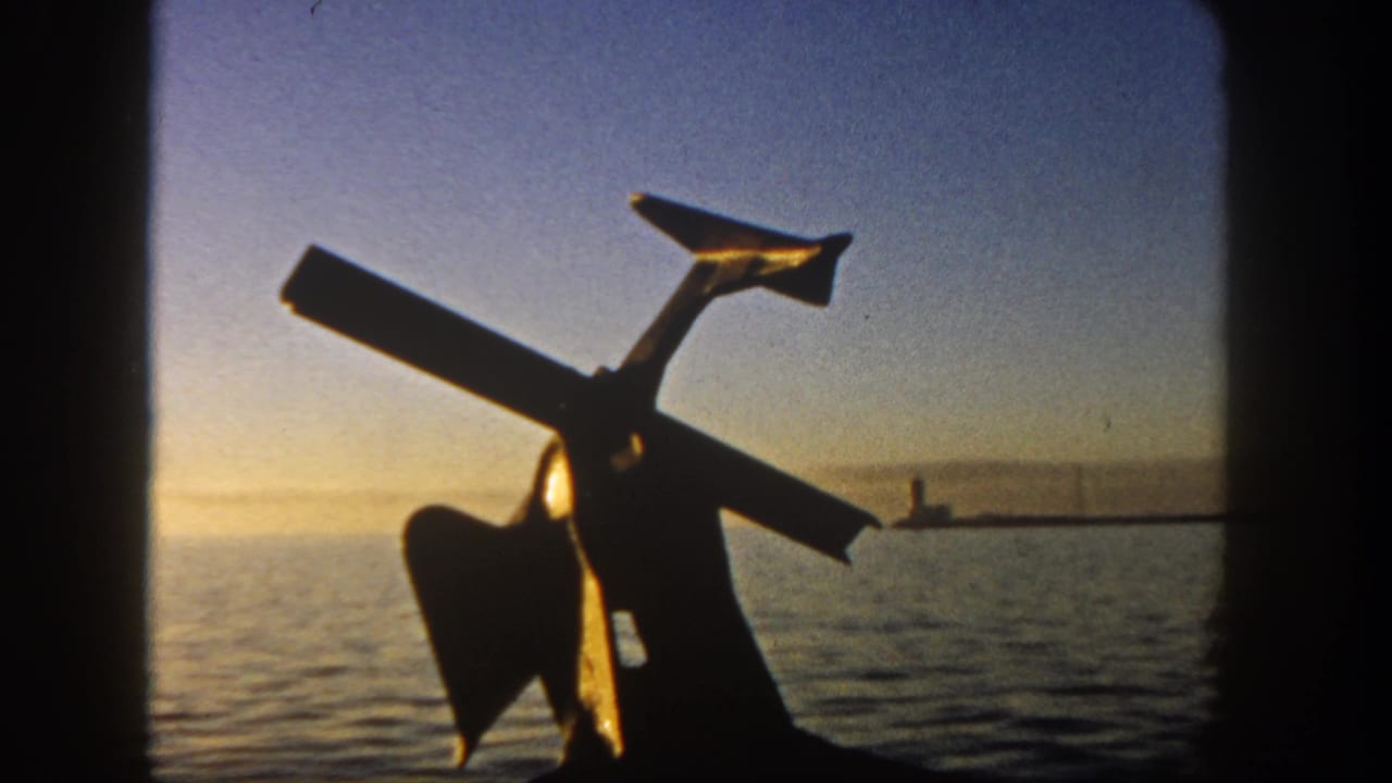 1962 San Diego 8mm frame showing a dark anchor-like form against calm harbor water and a distant lighthouse or pier marker at sunset.