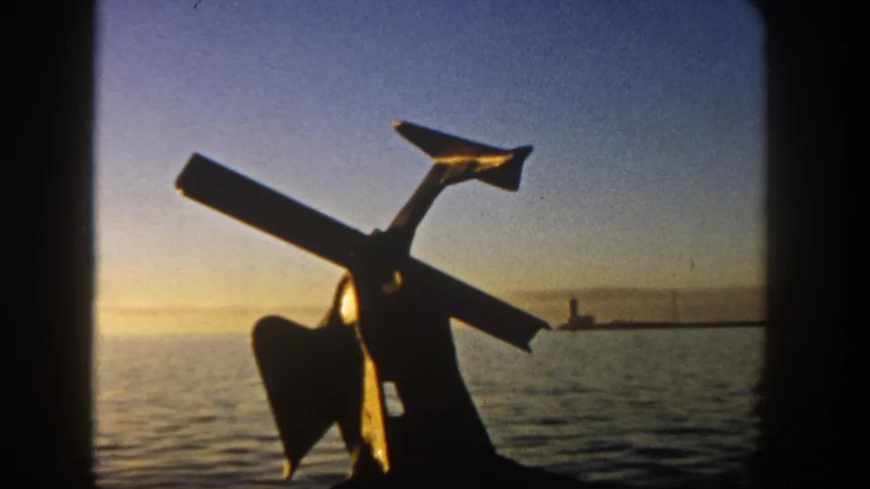 1962 San Diego 8mm frame showing a dark anchor-like form against calm harbor water and a distant lighthouse or pier marker at sunset.