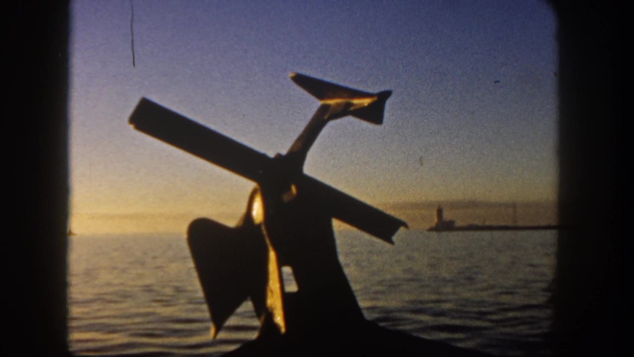 1962 San Diego harbor view on 8mm film, with an anchor-like silhouette, warm sky, rippled water, and distant pier structure.