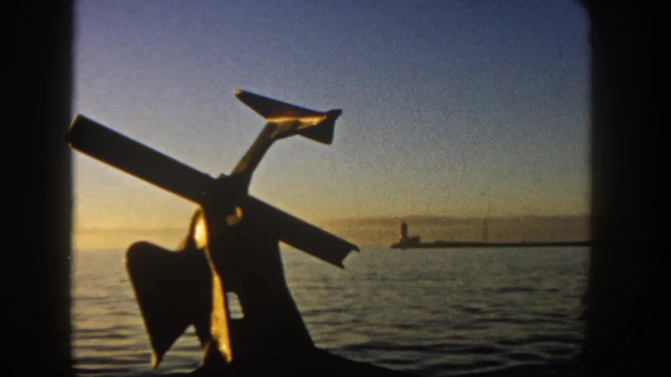 1962 San Diego waterfront footage frame with a dark anchor-like object, calm harbor water, sunset gradient sky, and distant pier light.