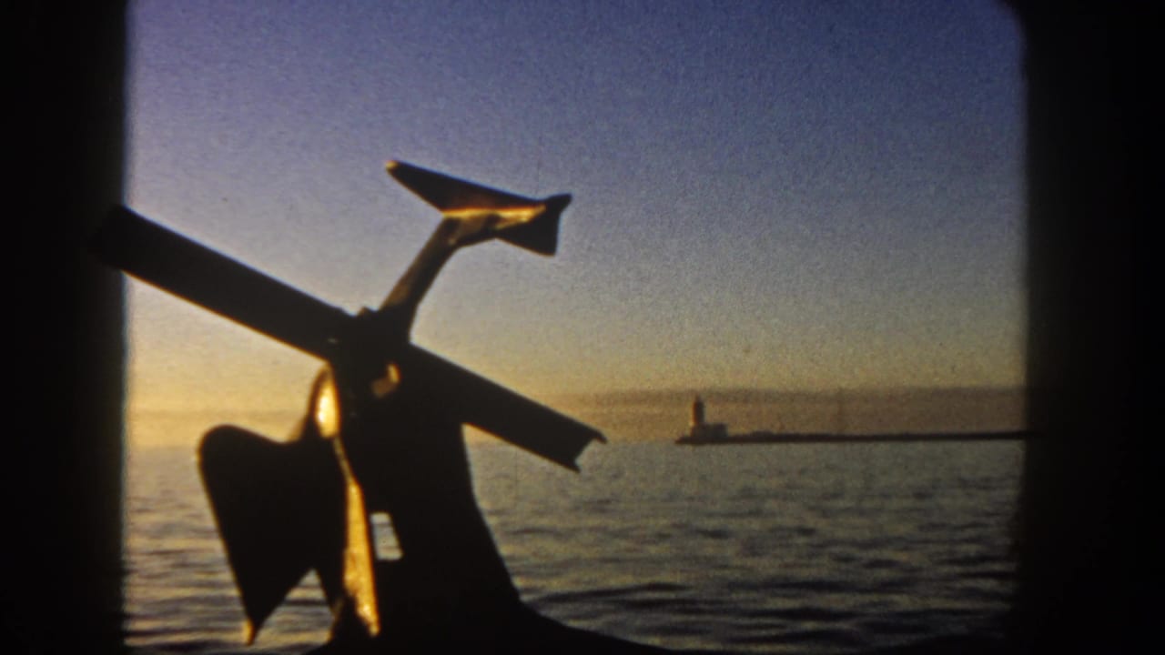 Silhouetted boat anchor in the foreground with calm water and a distant lighthouse near sunset.
