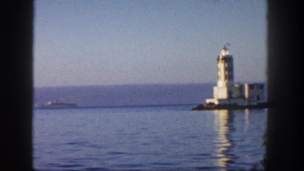 1962 San Diego coastal frame showing a white lighthouse and adjacent structures across still water, with its reflection visible.