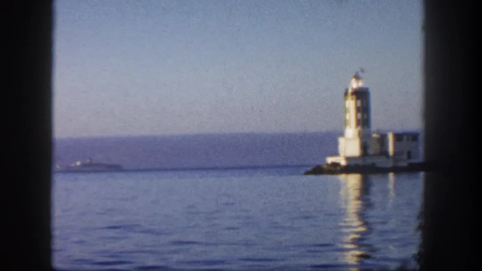 1962 San Diego coastal frame showing a white lighthouse and adjacent structures across still water, with its reflection visible.