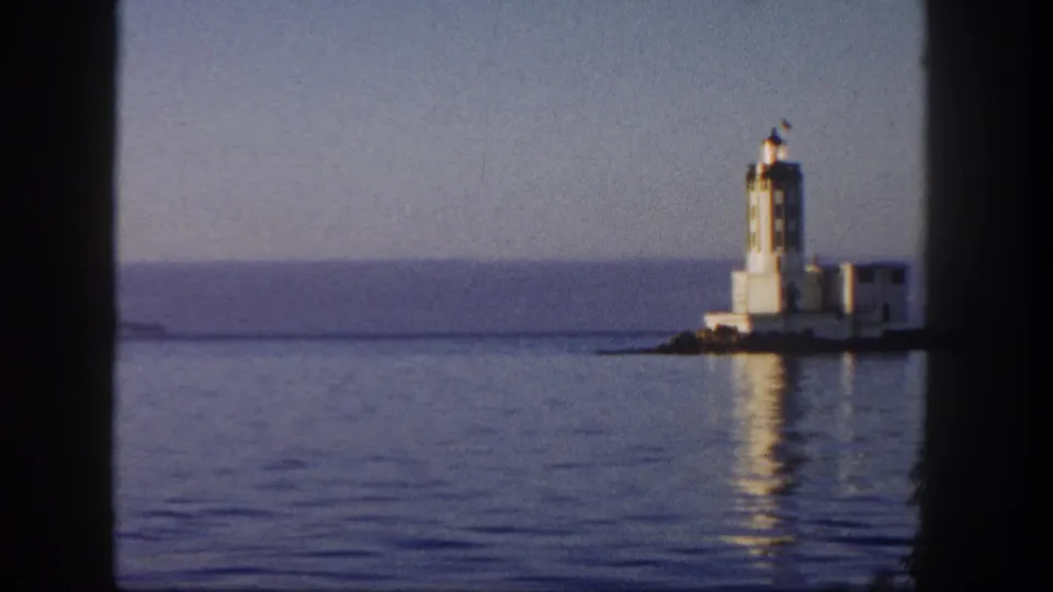 1962 San Diego boat-shot view with the lighthouse near frame edge, distant shoreline, smooth water, and a low breakwater or pier.