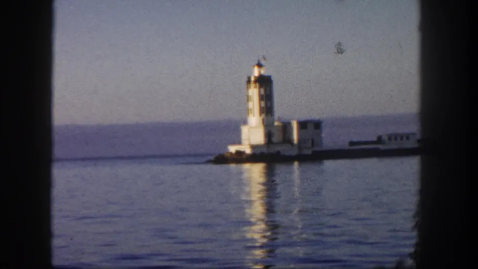 1962 San Diego coastal home-movie frame showing the lighthouse, nearby buildings, a pier or breakwater, and reflections on calm water.
