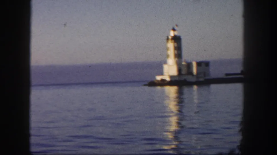 1962 San Diego 8mm frame of a lighthouse across calm water, partly framed by dark boat window mullions.
