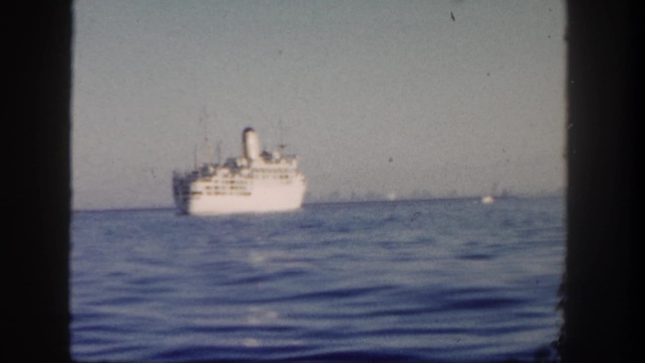 White passenger ship on calm blue water beneath a hazy sky, framed by dark film borders.