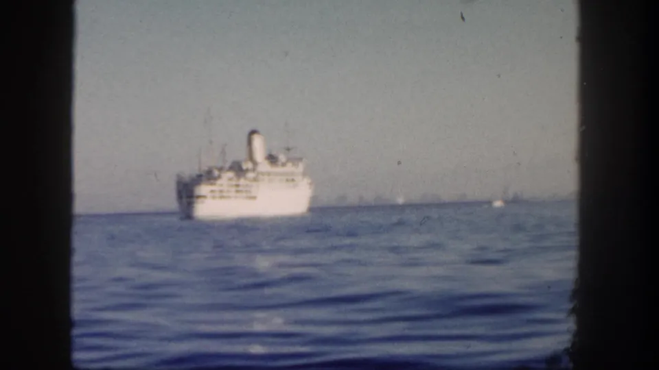 1962 San Diego home-movie frame showing a distant white passenger ship crossing calm blue water under a pale hazy sky.