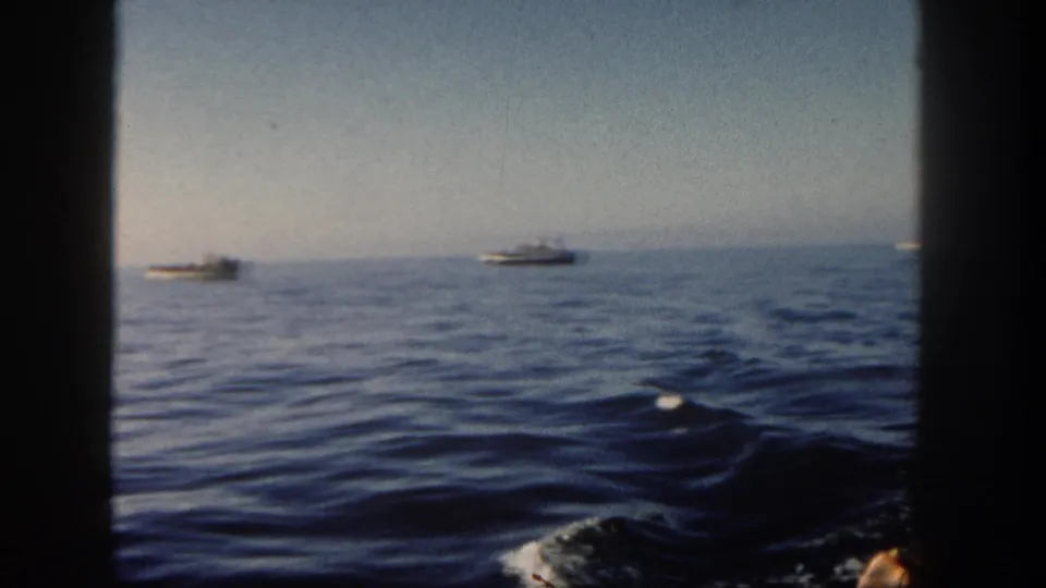 1962 San Diego ocean view from a moving vessel, with distant boats, low horizon, soft focus, and dark 8mm frame edges.
