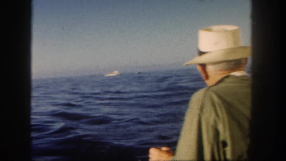 1962 San Diego home-movie frame with a passenger in a light brimmed hat facing the ocean and distant boats.