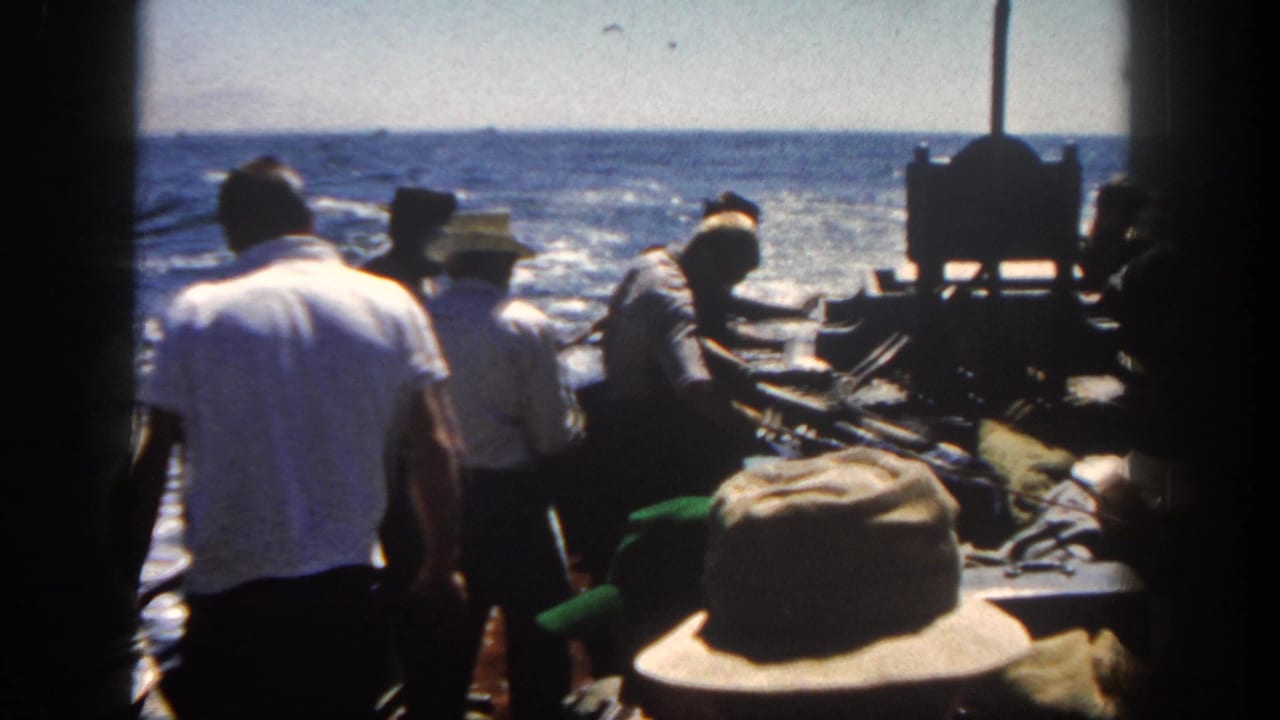 Adult men in casual 1960s clothing stand around gear on a moving boat off San Diego, with sunlit ocean behind them.