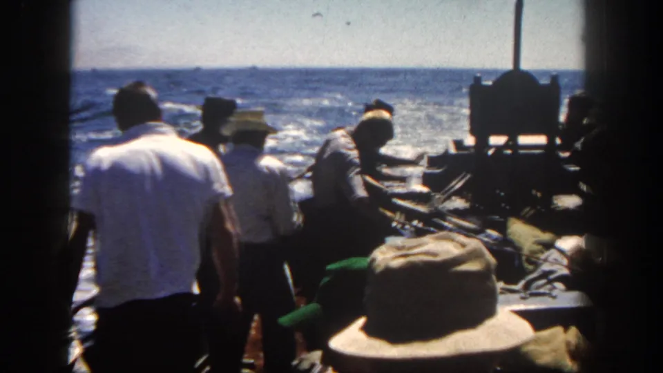 Adult men in casual 1960s clothing stand around gear on a moving boat off San Diego, with sunlit ocean behind them.