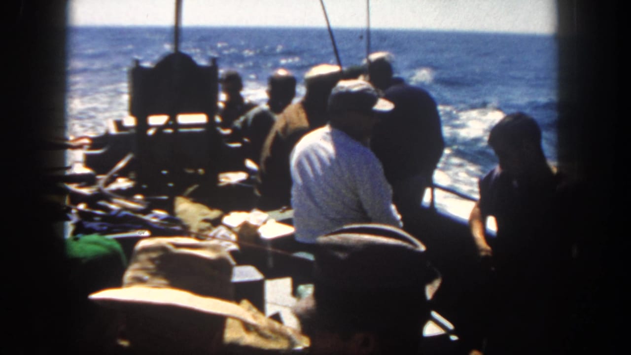 Several men face the stern area of a moving boat with ocean wake behind them.