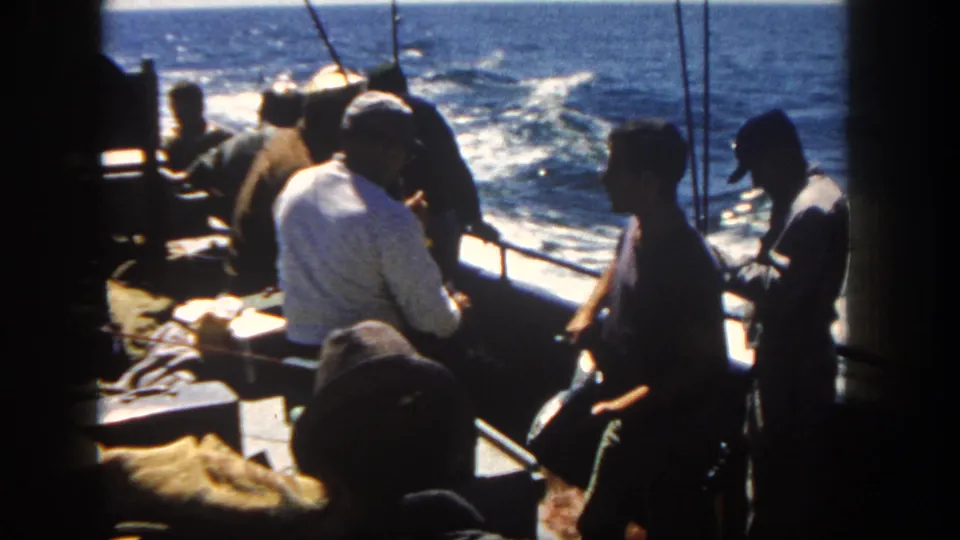 A man in a light shirt sits by the rail while others stand nearby on a 1962 San Diego fishing boat at sea.