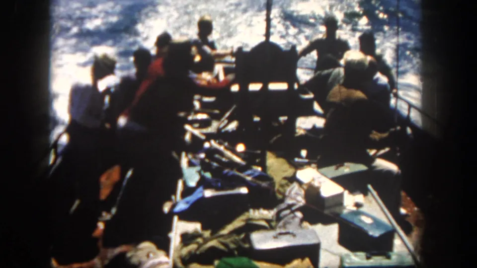 Backlit 8mm frame from 1962 San Diego shows men, rods, boxes, and deck gear on a fishing boat against sunlit water.