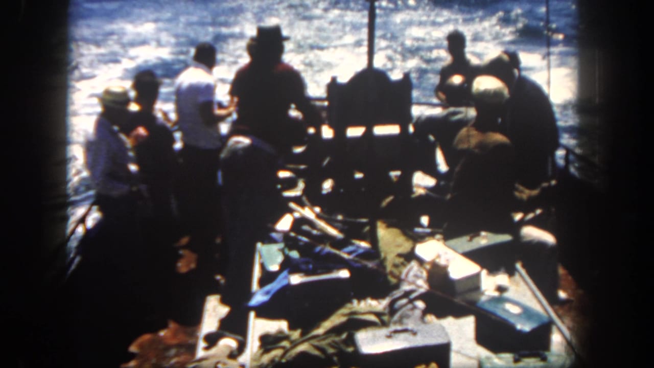 Men in hats and shirts stand around the stern of a boat with ocean wake behind them.