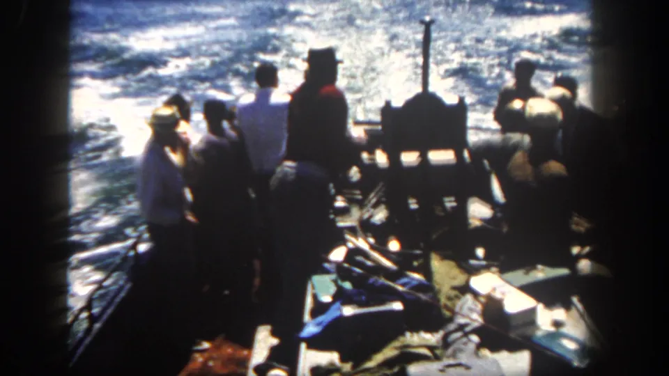 Silhouetted men remain grouped around the stern of a 1962 San Diego fishing boat as sun glare reflects across the surrounding ocean.