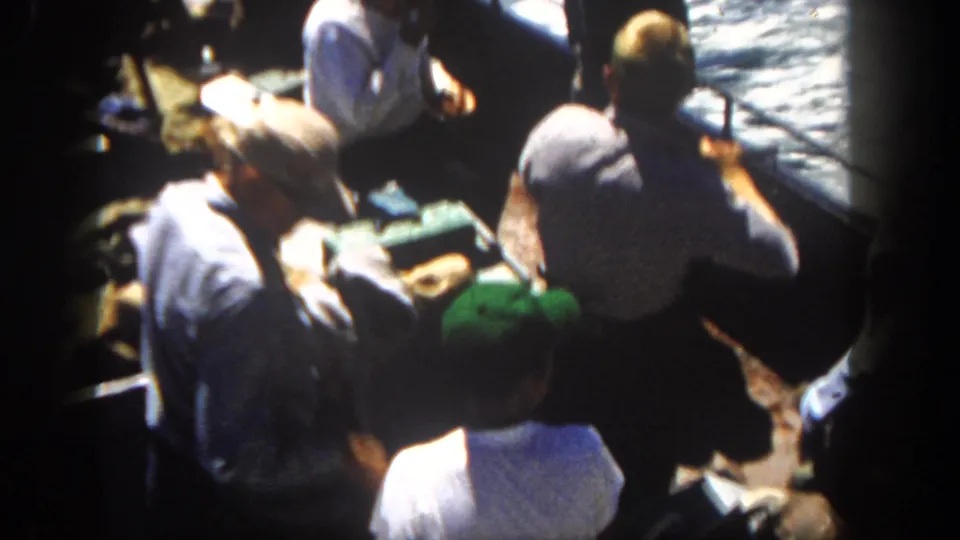 1962 San Diego 8mm frame with men in caps and shirts gathered around a tackle box on a crowded recreational boat deck.