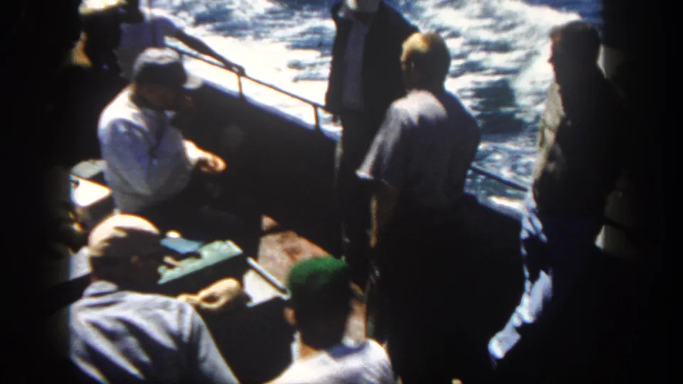 A 1962 San Diego home-movie frame shows men on deck near a railing with bright white wake and water alongside the boat.