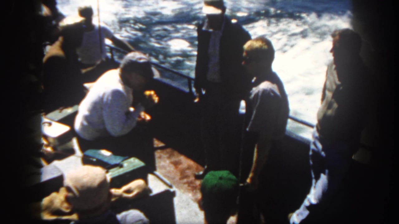 Passengers stand and sit on a boat deck beside churning water.