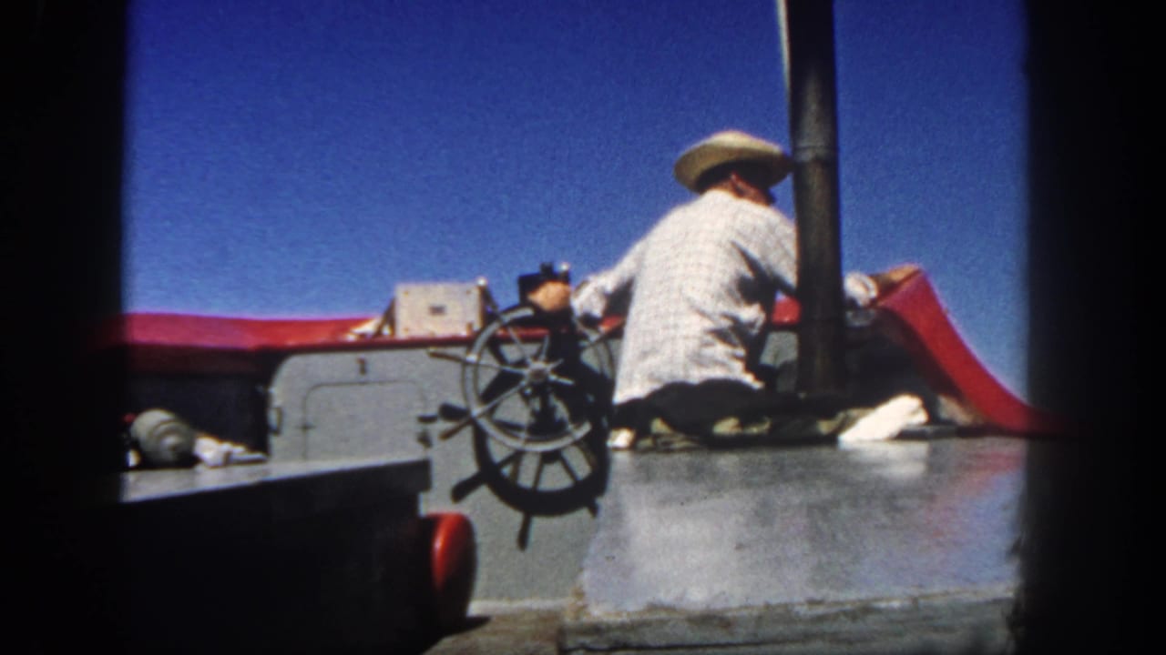 A man in a straw hat sits at a boat wheel under a blue sky.