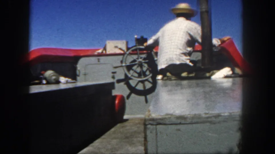 1962 San Diego 8mm frame showing a man in a straw hat steering from a boat deck under a blue sky.