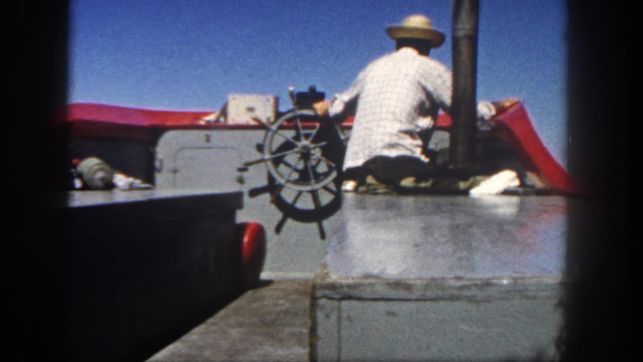 1962 San Diego home-movie frame of a man at a deck wheel, framed by dark film edges and bright red boat trim.