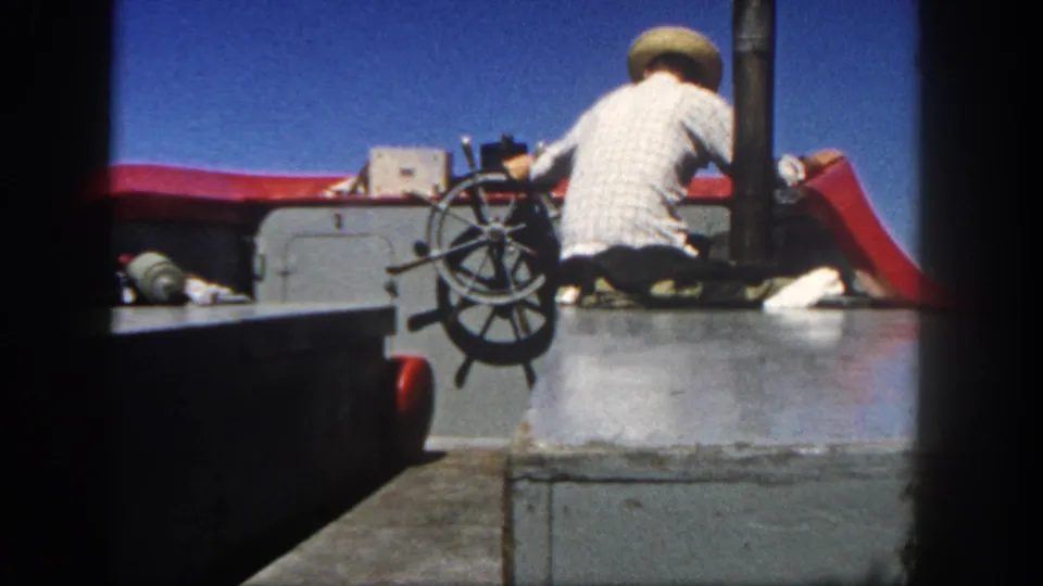 1962 San Diego 8mm frame with a closer rear view of a man in a straw hat handling the boat wheel.