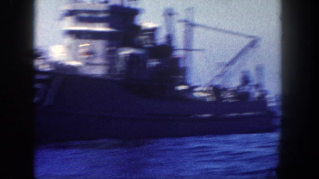 Side view of a large naval vessel on open water in 1962 home-movie footage, with deck structures, rigging, and distant crew shapes visible.