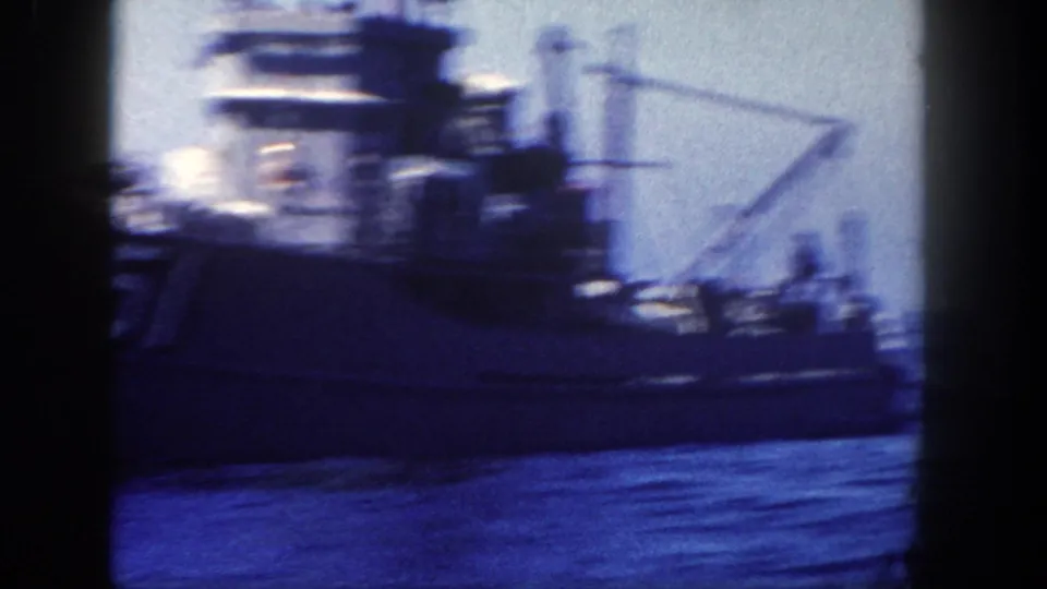 Side view of a large naval vessel on open water in 1962 home-movie footage, with deck structures, rigging, and distant crew shapes visible.
