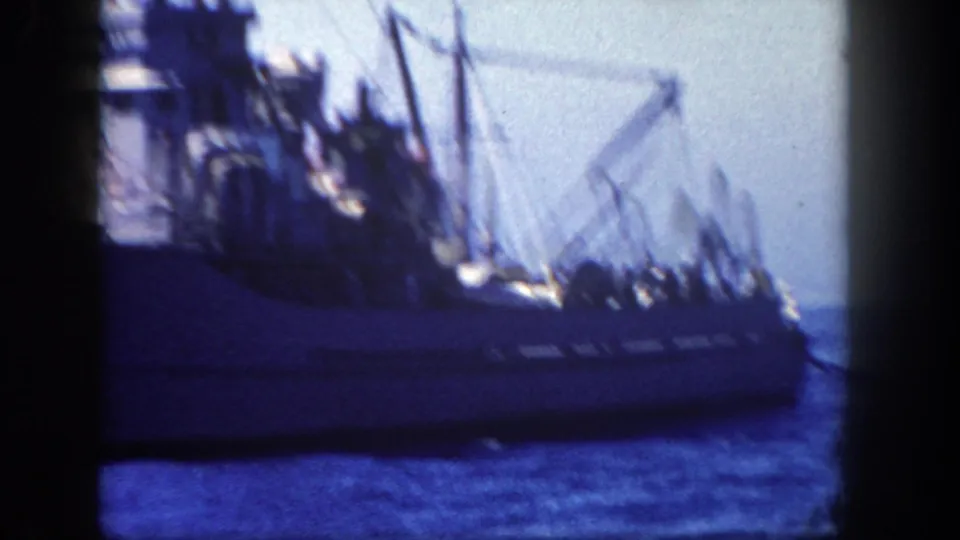 Blurred 8mm close view of the naval ship's deck area, showing rigging, equipment, and small crew silhouettes against bright water and sky.