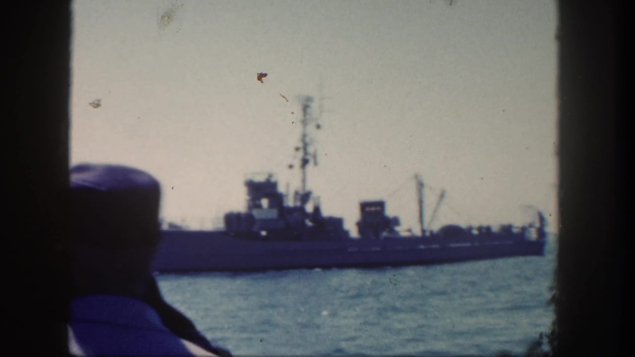 Sailor in foreground watches a naval ship on the water.